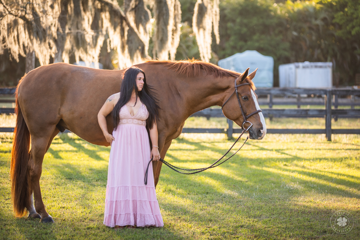 Photograph of a woman wearing a pink dress posed in front of a chestnut horse. They are both standing in front of backlight spanish moss dangling from a tree.