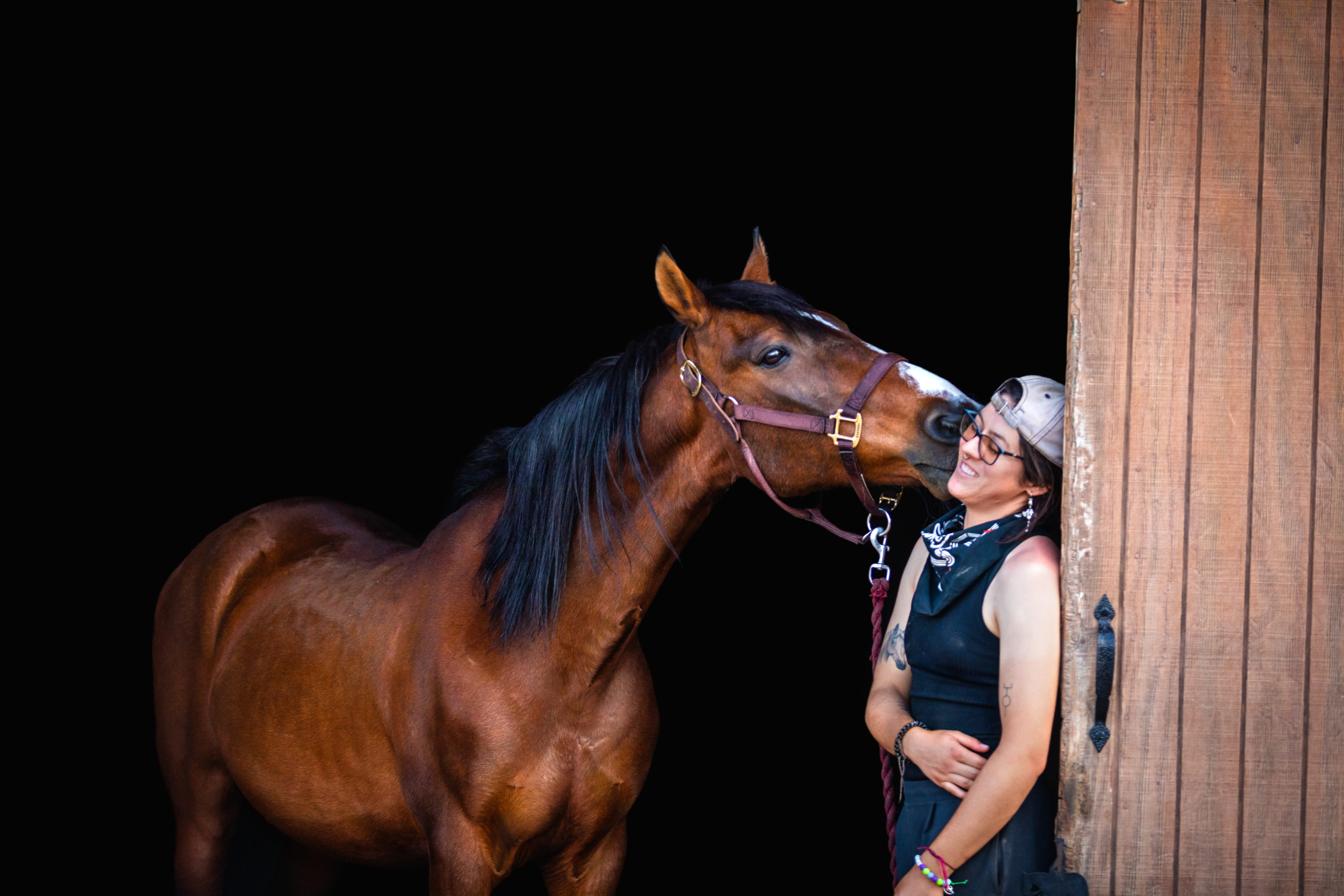 Photograph of a bay horse nuzzling a woman with short black hair against a black background.