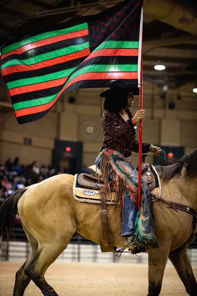 Photograph of a black cowgirl riding a horse with a black, green, and red American flag.