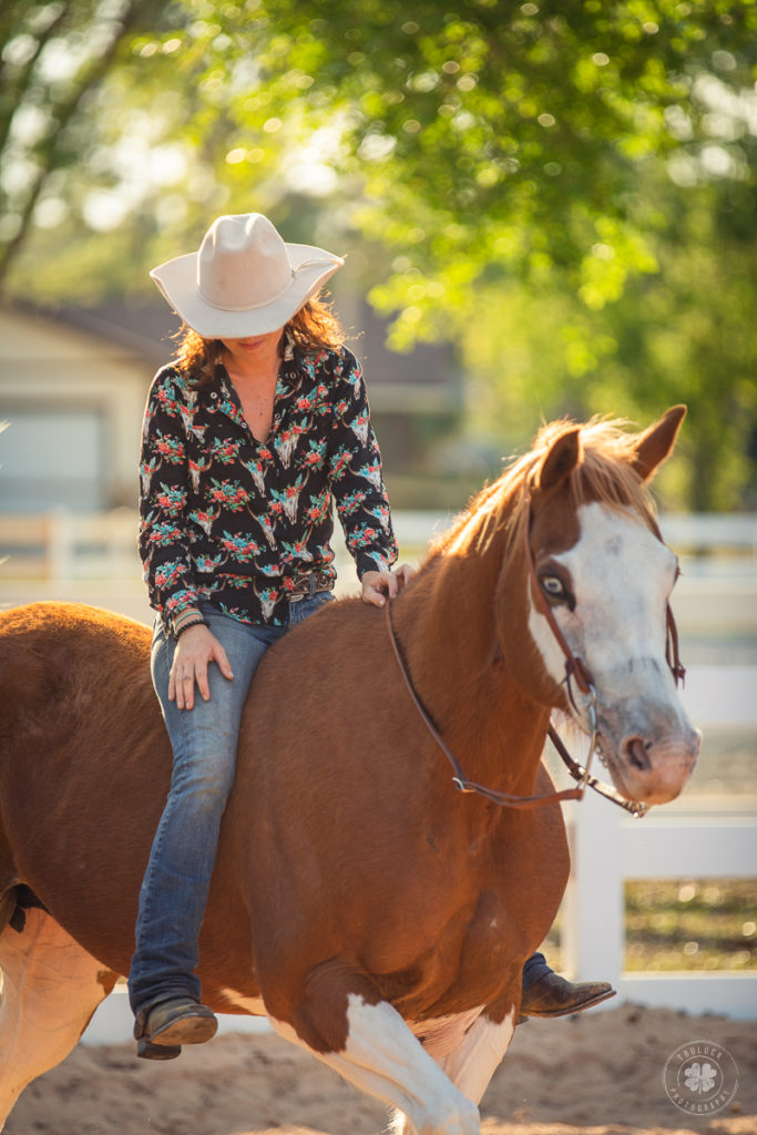 Photograph of a woman wearing a western floral cow skull shirt sitting on her horse.
