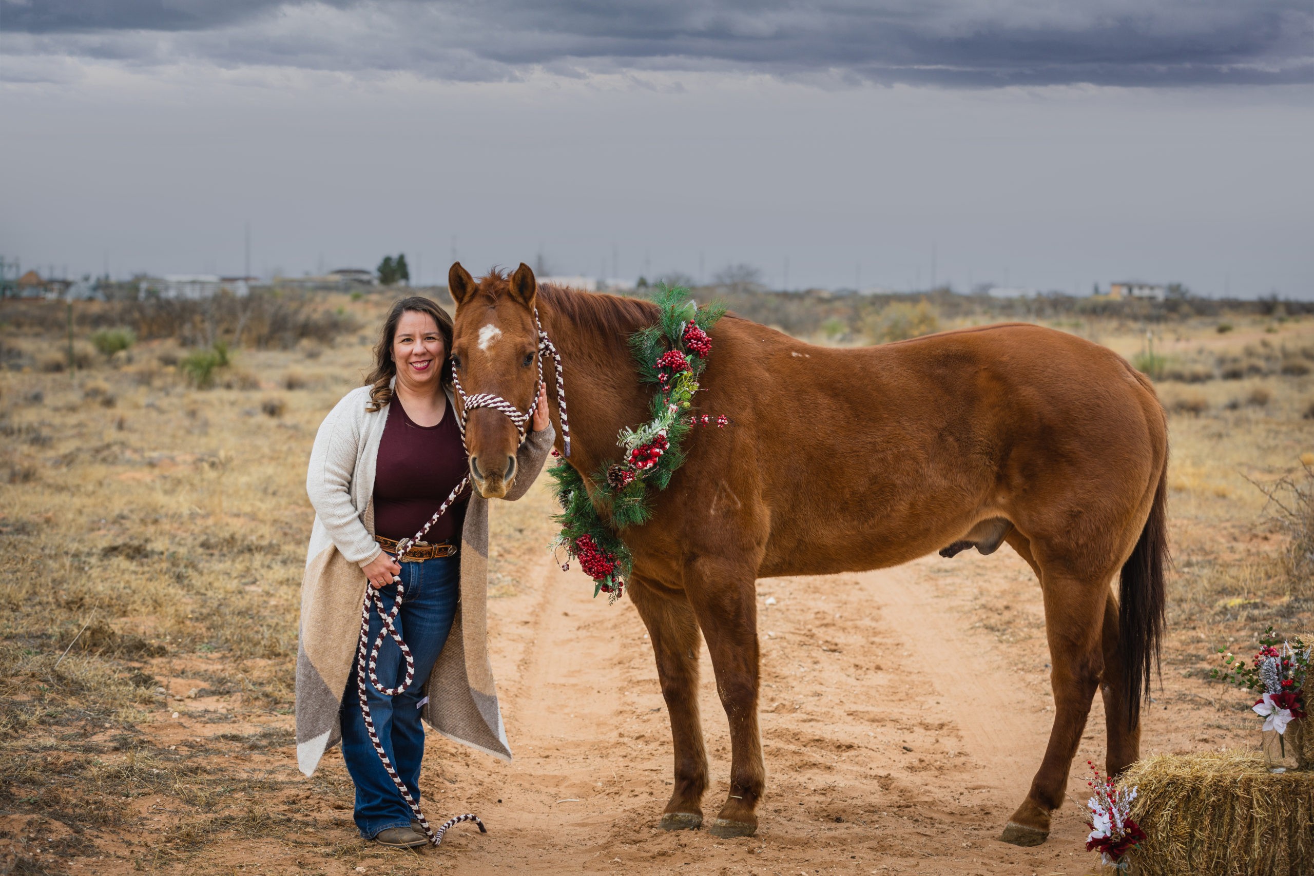Photograph of a woman posing next to a red dun horse wearing a Christmas wreath around his neck.