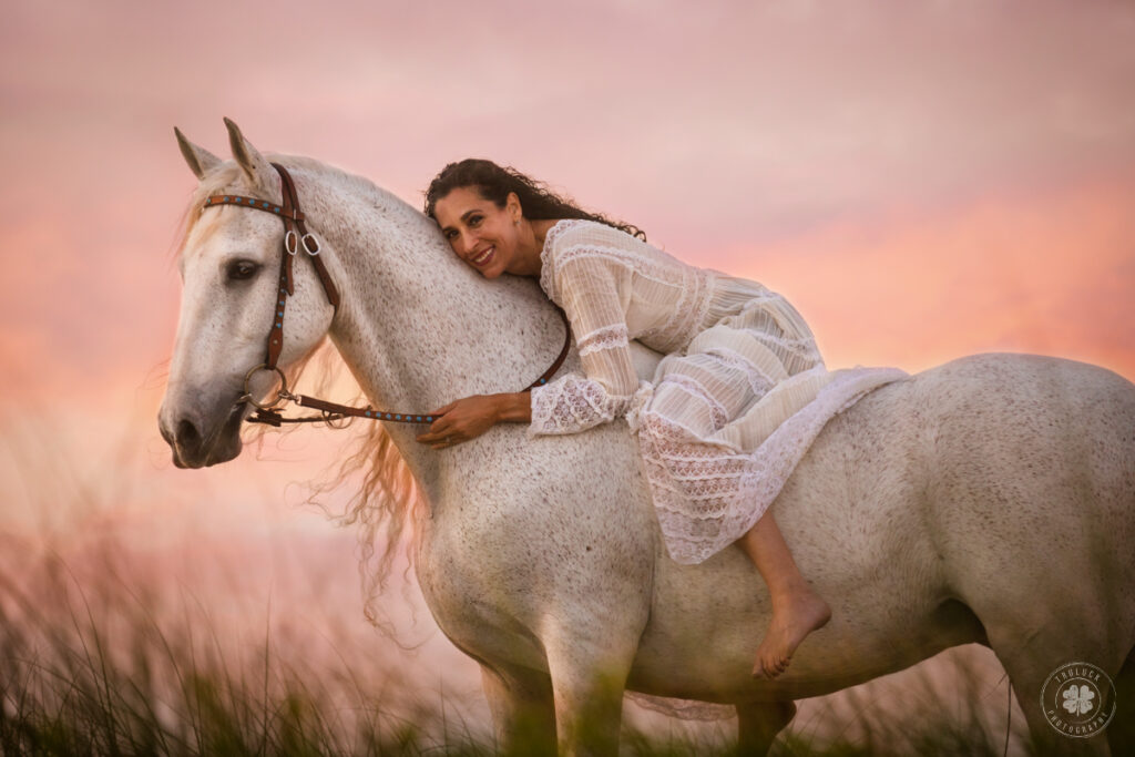 Photograph of a woman with long dark hair wearing a white dress sitting bareback on a white horse.  