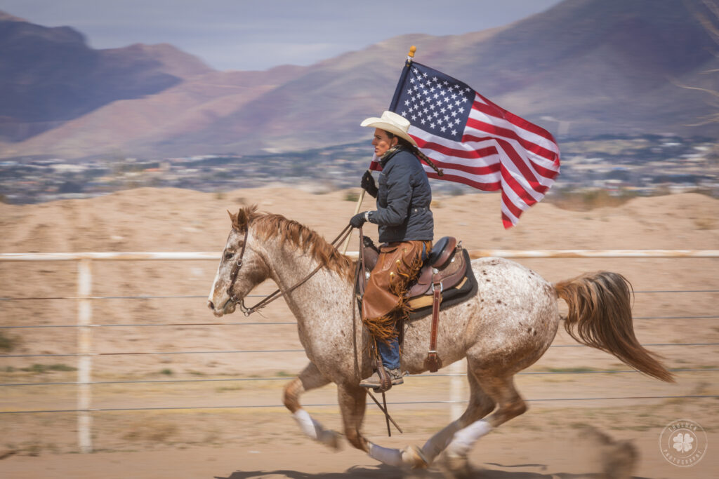 Photograph of a cowgirl galloping a horse around the arena while carrying an American flag. 
