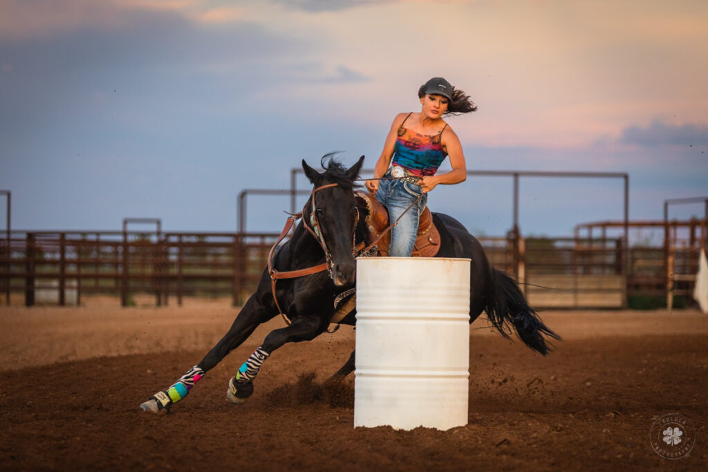 Photograph of a cowgirl riding a black horse around a white barrel in an arena in Deming, New Mexico. 