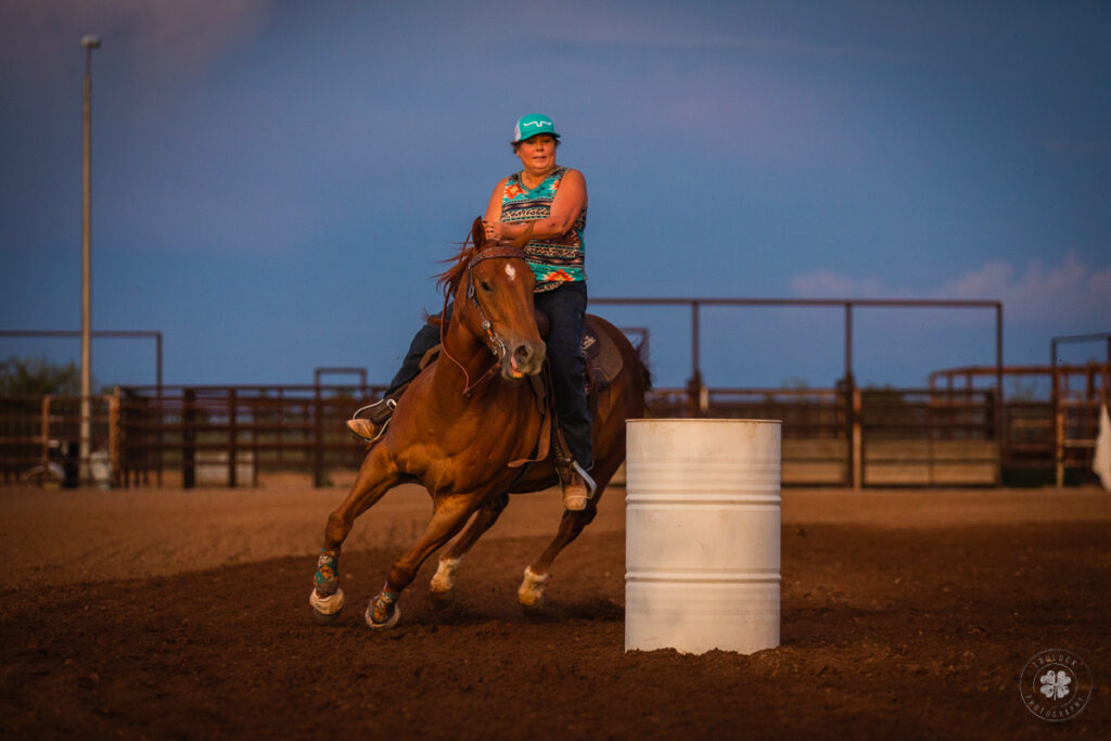 Photograph of a cowgirl riding a chestnut horse around a white barrel during a barrel race in Deming, New Mexico. 