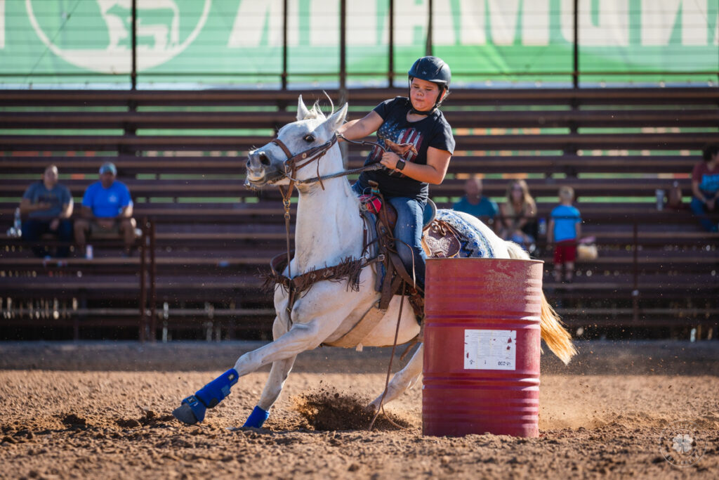 Photograph of a cowgirl wearing a helmet riding a gray horse around a barrel during a barrel race in Mesquite, New Mexico. 