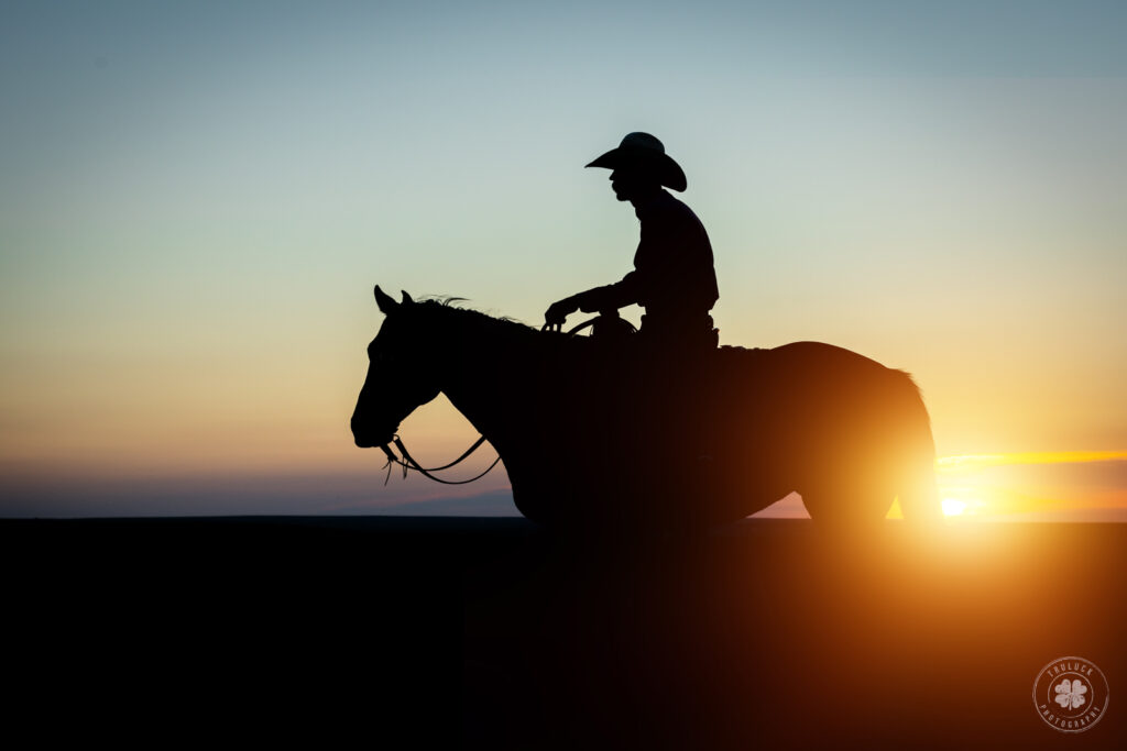 A silhouette of a cowboy on his horse with the sun shining near the horse's tail. 
