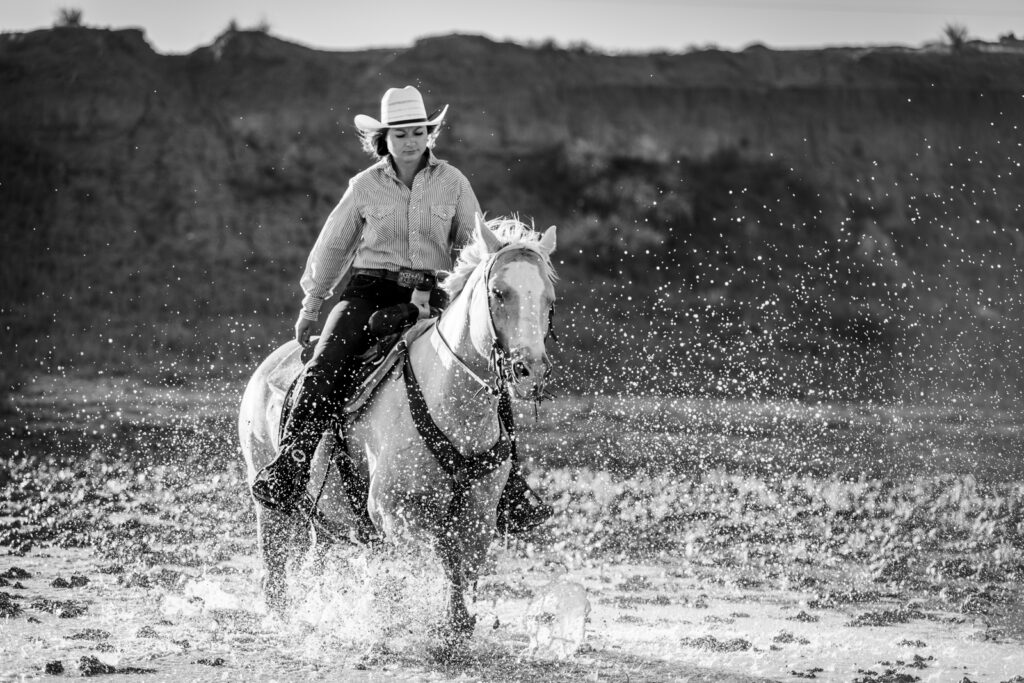 A black and white photograph of a cowgirl riding her horse through water.  