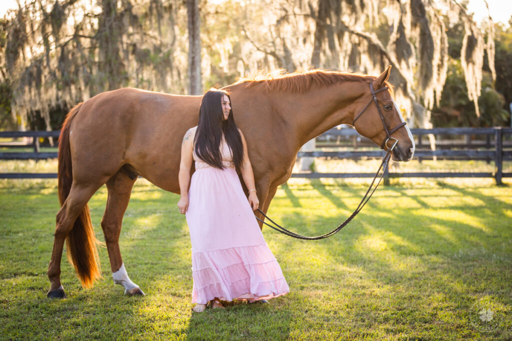 Photograph of a woman with long dark hair wearing a pink dress standing in front of a chestnut horse in a field with trees in the background near Bradenton, Florida. 