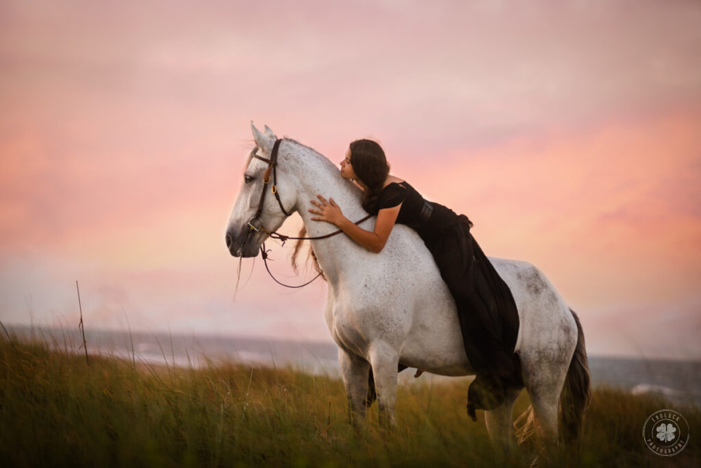 Photograph of a girl wearing a black dress laying bareback on her gray horse against the backdrop of a sunset on a beach in Florida.  