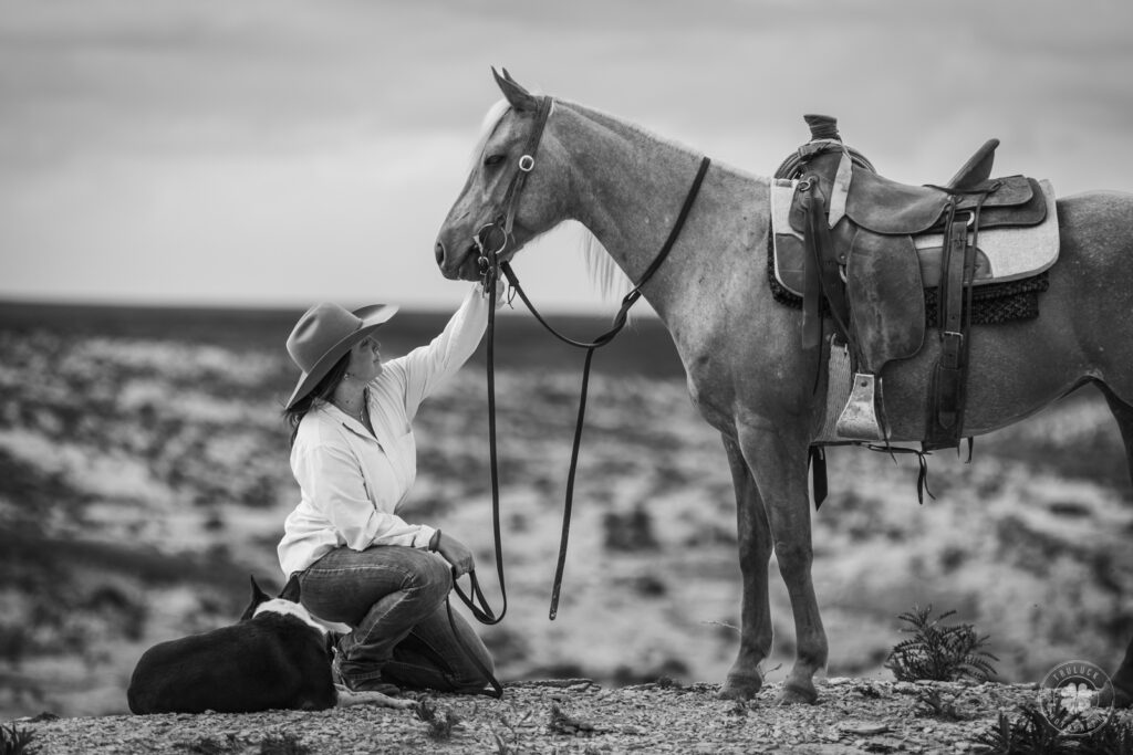 Black and white photograph of a cowgirl kneeling in front of a horse with her arm outstretched to pet her nose in Scott City, Kansas. 