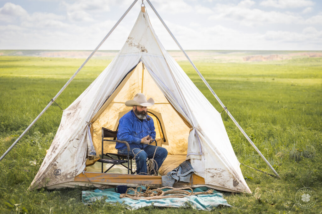 Photograph of a cowboy braiding a hackamore inside of a cream colored tent.  