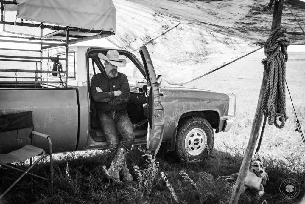 Black and white photograph of a cowboy leaning against the side of his truck with his arms and legs crossed.  