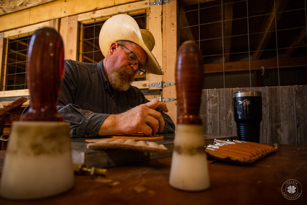 Photograph of a cowboy carving leather while framed by some of his tools.  