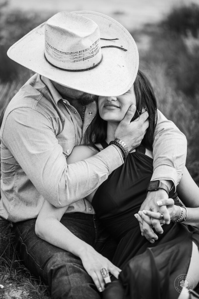Black and white photograph of a couple.  The woman is leaning against her husband and his arms are wrapped around her.  His head is tilted so that his cowboy hat covers her eyes.  