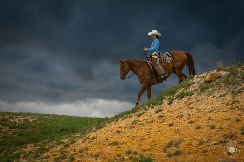Photograph of a cowgirl riding her horse down a steep hill in front of a dark thunderstorm in Kansas.  