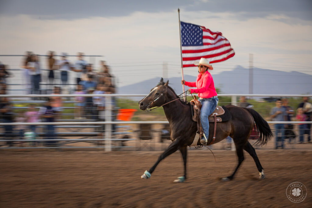 A panned photograph of a cowgirl riding a dark bay horse while carrying the American flag during a ranch rodeo in Deming, New Mexico. 