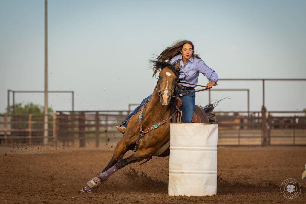 Photograph of a cowgirl riding her horse around a white barrel during a rodeo in Deming, New Mexico. 