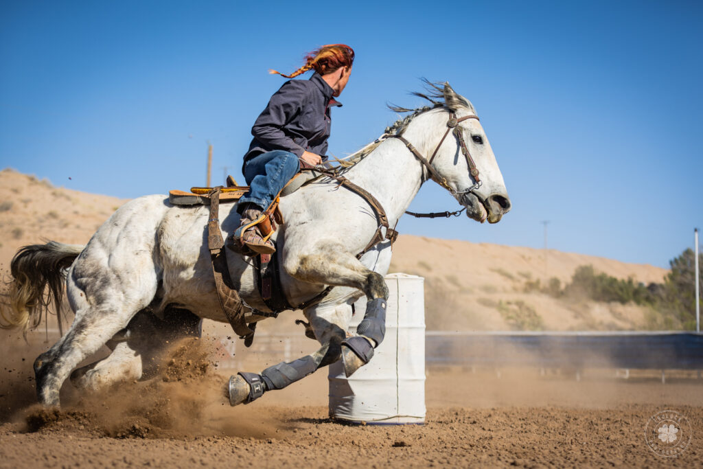 A low angle photograph of a cowgirl running a gray horse around a white barrel in west Texas. 
