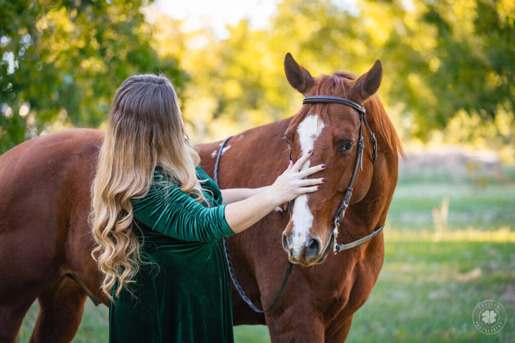 Photograph of a blonde woman wearing a green dress petting the nose of a chestnut horse in an orchard in Las Cruces, New Mexico. 