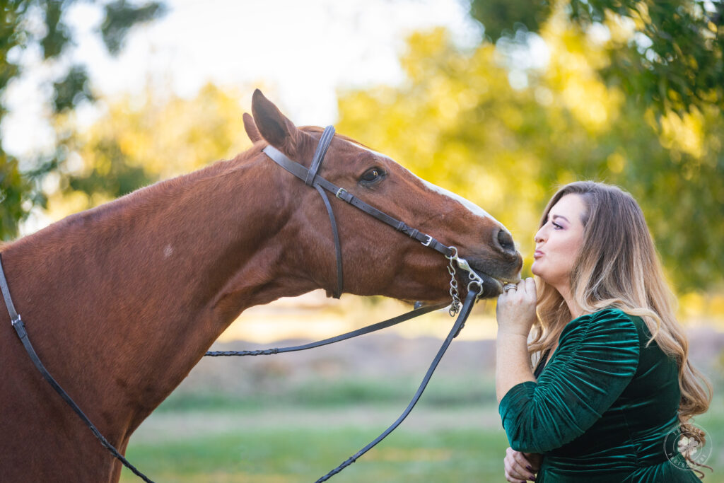 Photograph of a woman wearing a green dress kissing the nose of a chestnut horse. 