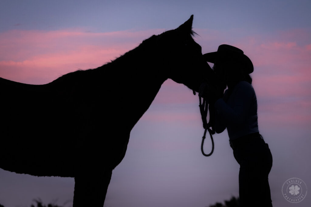 Silhouette of a woman wearing a cowboy hat kissing her senior horse on the face. 