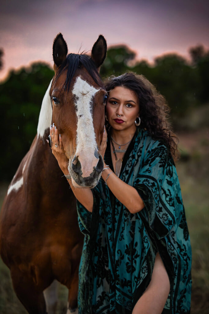 Photograph of a woman wearing a teal and black kimono with her hands on the cheeks of a brown and white paint horse.  They are both facing the camera. 