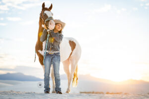 Photograph of a teenager wearing jeans and a denim jacket standing in front of a brown and white paint horse on white sand dunes in White Sands National Park. 