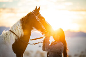 Photograph of a teenage girl resting her head against her horse's muzzle.  The sun is bursting between their heads. 