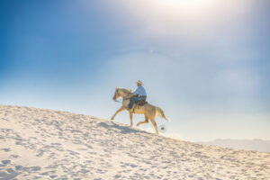 Photograph of a cowboy riding a palomino horse up a white sand dune against a blue sky. 