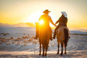 Photograph of a couple holding hands looking at the sunset while riding horses at White Sands National Park.