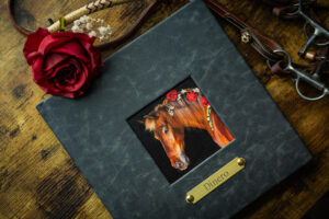 A photograph of a black leather album with a square cutout showing a photograph of a horse's head.  There is a halter nameplate under the photo with the name "Dinero" inscribed underneath it.