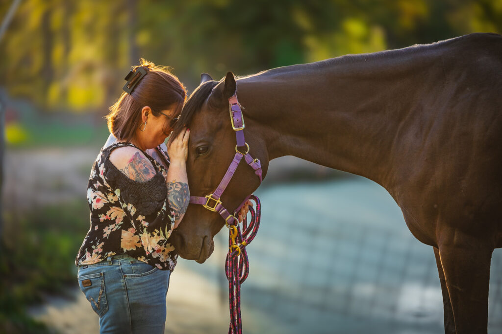 Photograph of a woman wearing a black floral shirt and jeans leaning her head on the head of a bay Thoroughbred mare in Clarksville, Tennessee.