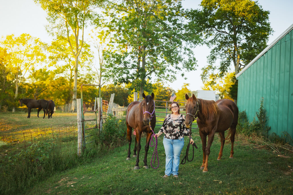 Photograph of a woman wearing a floral black shirt and jeans standing between two Thoroughbred mares in a field beside a green barn.