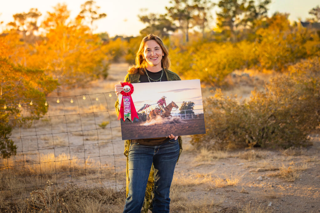Photograph of a woman in jeans and a green duster holding a metal print of a cowboy spinning his horse in the dust while holding an American flag on a pole behind him in El Paso, Texas.
