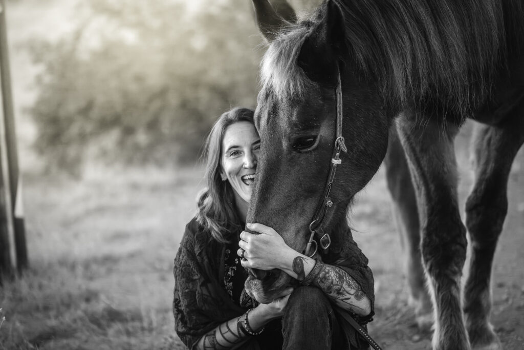 Black-and-white-photograph of Kristina laughing at the camera as she hugs her horse's head while sitting on the ground in El Paso, Texas.