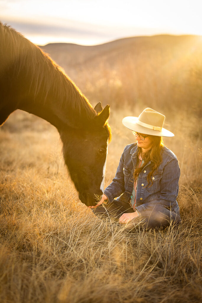 Photograph of a horse sniffing her owner's hand as she sits cross-legged in a golden field in Las Cruces, New Mexico with a hill in the background. She is wearing a jean jacket and a tan fedora.