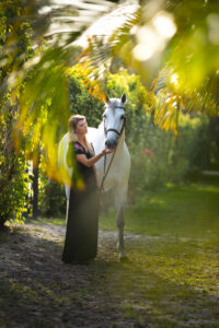 A woman in a black dress stands next to a gray horse, holding her hand to its muzzle. They are framed by palm fronds.