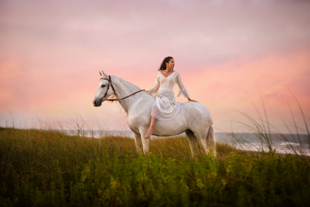 Photograph of a woman sitting on a gray horse looking behind her against a brilliant pink and lavender sunset. 