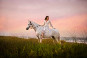 Photograph of a woman sitting on a gray horse looking behind her against a brilliant pink and lavender sunset.