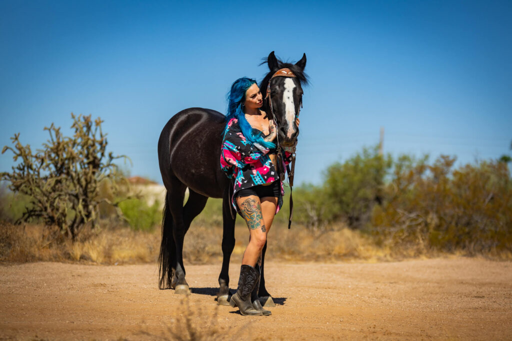 A punk rock girl with blue hair and bold blue, pink, and black clothes poses next to her black mustang in the afternoon sun in the desert. 