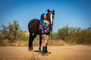 A punk rock girl with blue hair and bold blue, pink, and black clothes poses next to her black mustang in the afternoon sun in the desert.