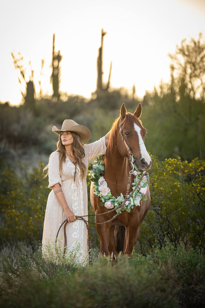 Photograph of a cowgirl wearing a cream dress standing next to a chestnut horse wearing a pink flower garland in the southwestern desert. 