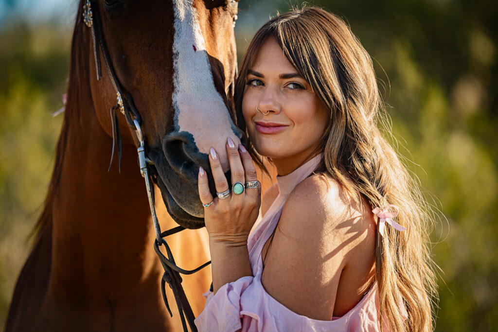 Closeup portrait of a woman in an off-the-shoulder pink top with her hand on the muzzle of a chestnut horse with a blaze in the desert. 