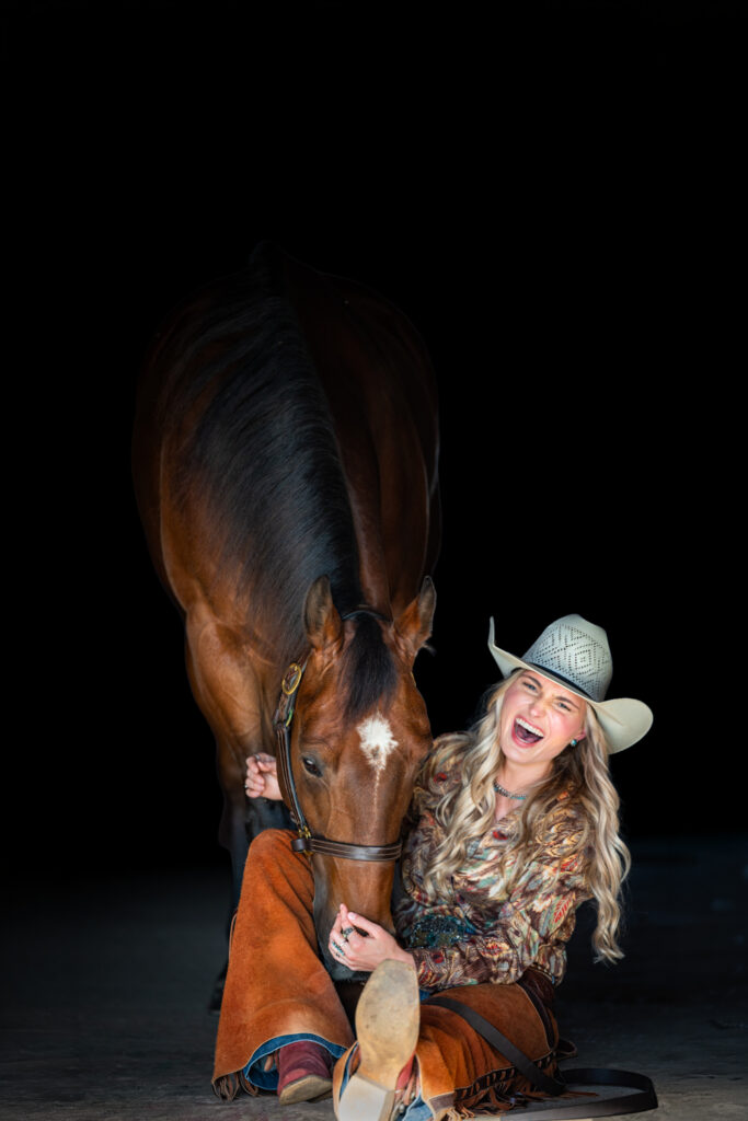 Photograph of a college girl sitting on the floor laughing as her bay horse lowers his head to nuzzle her for a black background photograph.