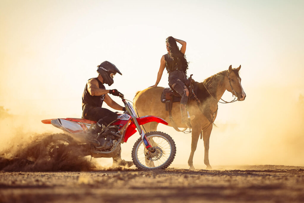 Photograph of a man riding a dirt bike next to a woman posing on a buckskin horse against a dusty background in the desert