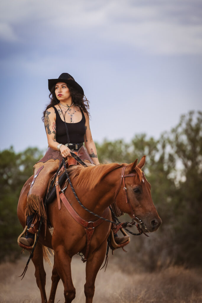 Photograph of a woman riding a chestnut horse wearing a black tank top, tan chaps, and a black cowboy hat in El Paso, Texas.
