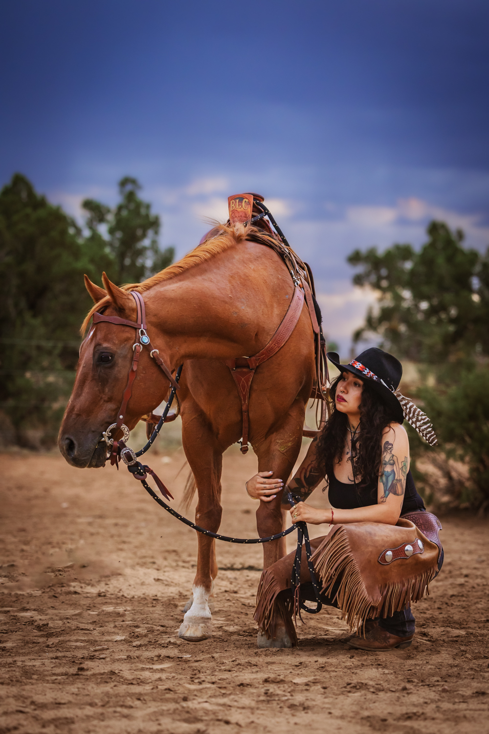 Photograph of a Latina woman wearing a black tank top and tan chaps kneeling next to her chestnut horse in El Paso, Texas.