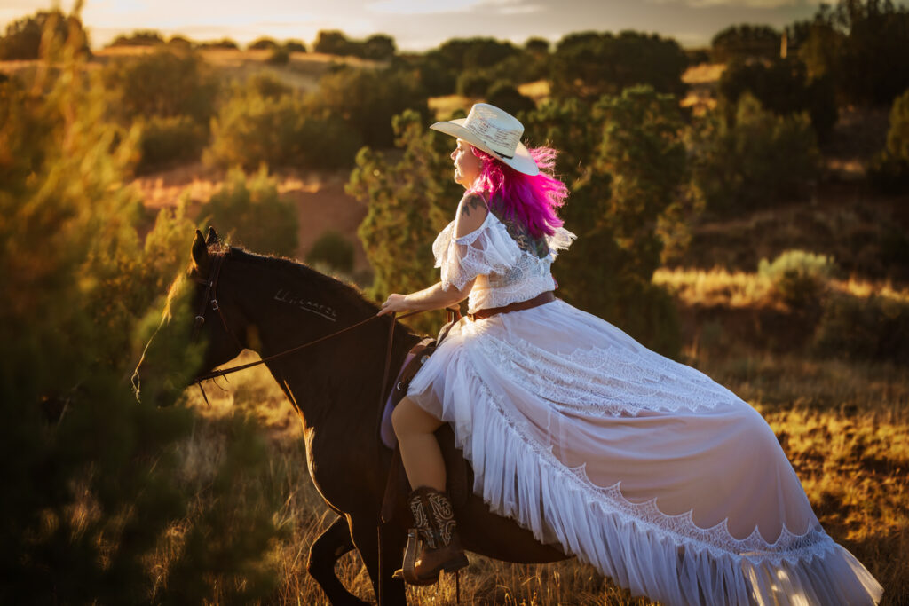 Photograph of a woman with pink hair wearing a cowboy hat and a long white dress riding her dark bay Mustang gelding.